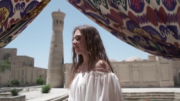 在一座古老的尖塔旁边的广场上 站着一位年轻女子 她的头发飘扬 身穿浅白色的太阳衣 乌兹别克斯坦慢动作 — 图库视频影像