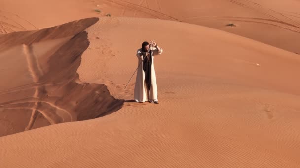 砂漠の砂の上で踊るシャーマンの格好をした若い髭の男 ドローン航空 — ストック動画
