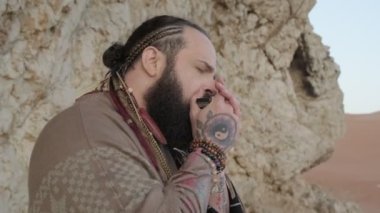 Şaman gibi giyinmiş bir adam çölün ortasında bir kayanın üzerinde duruyor ve mızıka çalıyor. Ağır çekimde kapat