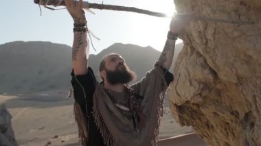 Elinde asası olan genç sakallı bir adam çölün kumlu kumları arasında bir kayanın üzerinde dua ediyor. Ağır çekim, BAE