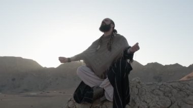 Lotus pozisyonunda genç bir şaman çölün kum tepeleri arasında gün batımının arka planına karşı bir kayanın üzerinde dua eder. Ağır çekim, BAE