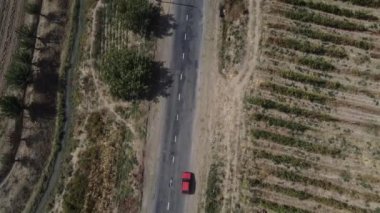 İnsansız hava aracı bozkır ve çalılarla çevrili bir kır yolunun üzerinde uçuyor. Yol boyunca farklı renklerde arabalar sürüyor. Hava görünümü