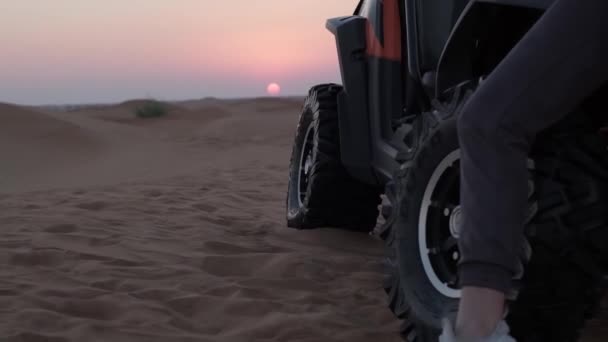 スポーツウェアに身を包んだ若い女性は 砂漠の砂丘で夕日を背景にバギーの横に立っている スローモーション — ストック動画