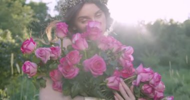 A young girl with a wreath of flowers holds a large bouquet of roses among the spring garden. Slow motion