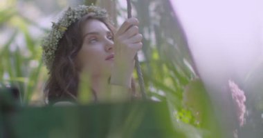A young beautiful girl in a wreath of flowers swings on a swing among a green garden. Slow motion
