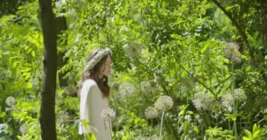 A young woman with a wreath of flowers on her head walks in a white light dress in the garden among green leaves and flowering plants. slow motion