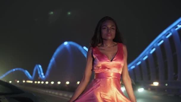 長い夜のドレスを着たスワロシーの若い女性は ライトアップされた夜の橋を渡ってスポーツカーに向かいます スローモーションドバイ — ストック動画