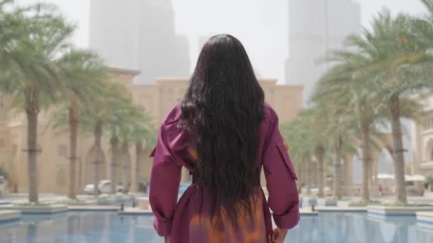 長い黒い髪と短い色のマントを持つ若い女性が噴水の端に沿ってカメラから歩いています スローモーション アラブ首長国連邦 — ストック動画