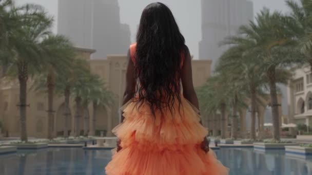 長いピンクのドレスを着た黒い髪の若い女性が噴水の端に沿ってカメラから歩いています スローモーション ドバイ アラブ首長国連邦 — ストック動画