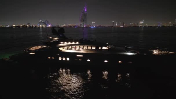 ドローンは ドバイの高層ビルの背景に夜に立っている照らされた高価なプライベートヨットの周りを飛び回ります エアリアルビュー スローモーション — ストック動画