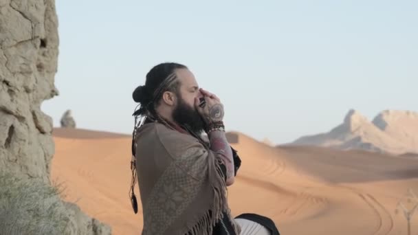 男はシャーマンの格好をし 砂漠の砂の真ん中の岩の上に立ってハーモニカを演奏した 砂漠Uae スローモーション — ストック動画