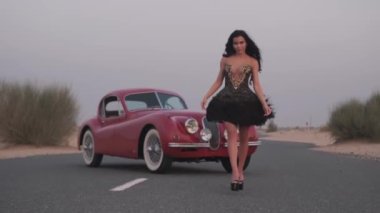 Siyah elbiseli, uzun saçlı ve parlak makyajlı genç bir kadın. Kırmızı bir retro arabanın yüksek topuklu ayakkabılarıyla yolda yürüyor. Yavaş Hareket