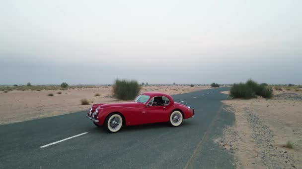 無人機は砂漠の真ん中に空の道に立っている高価な赤いヴィンテージ車の周りを飛ぶ エアリアルビュー スローモーション — ストック動画