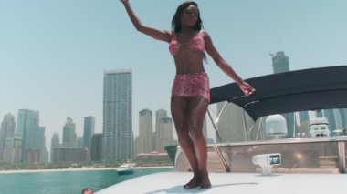 Özel bir teknenin güvertesinde Dubai gökdelenlerinin önünde dans eden kısa parlak elbiseli esmer bir kadın. BAE, Ağır çekim