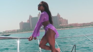 Beyaz bikinili esmer bir kadın ve şehrin arka planına karşı özel bir teknede açık renk bir elbise giyiyor. Ağır çekim, BAE