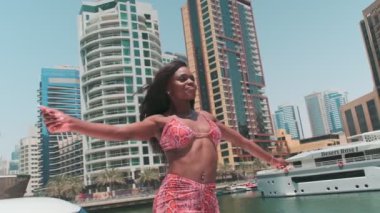 Dubai gökdelenlerinin arka planına karşı özel bir teknenin güvertesinde kısa parlak elbiseli esmer bir kadın. BAE, Ağır çekim