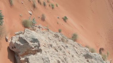 Bir dron, kumlu çölün ortasında, ıssız bir kayanın üzerinde duran kırmızı elbiseli genç bir kadına doğru uçuyor. Hava görünümü.