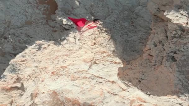 ドローナは 砂漠の真ん中に崖の上に立っている明るい赤いドレスで若い女性の上を飛ぶ エアリアルビュー — ストック動画