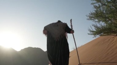 Beyaz şaman elbiseli genç bir adam elinde asasıyla çölün kumlarında gün batımına karşı yürür. Yavaş çekim BAE