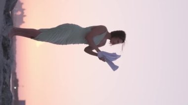 Hafif elbiseli genç bir kadın günbatımında denizin arka planında duran bir kayanın üzerinde rüzgarda kağıtları savuruyor. Dikey, Yavaş Hareket