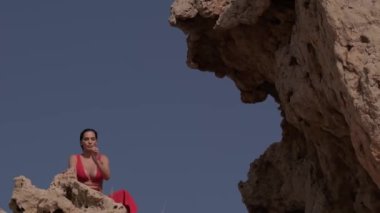 Kırmızı elbiseli genç bir kadın mavi gökyüzüne karşı çölün ortasında bir kayanın üzerinde oturuyor. Yavaş Hareket.