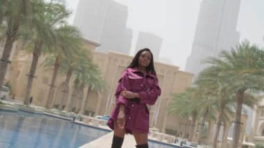 Kısa pelerinli ve yüksek çizmeli güzel bir kadın parkta açık bir fıskiyenin yanında duruyor. Yavaş Hareket, BAE