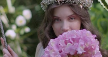 A young beautiful girl in a wreath of flowers swings on a swing among a green garden. Slow motion