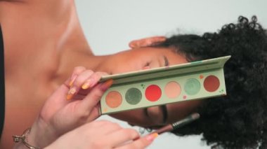 Profesyonel makyaj ustası kıvırcık saçlı genç bir modele boya uyguluyor. Dikey, Yavaş Hareket, Kapat.