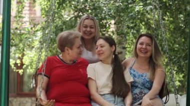 Güneşli bir günde meyve bahçesinde salıncakta sallanan üç kızı olan yetişkin bir kadın. Sarılırlar ve gülerler. Yavaş Hareket.
