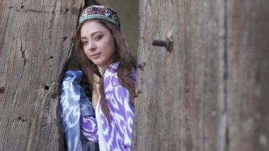 Uzun saçlı, kısa beyaz elbiseli ve ahşap bir kapının arkasında oryantal cüppeli genç bir kadın. Yavaş Hareket, Özbekistan