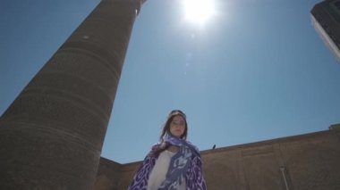 Beyaz elbiseli ve uzun doğulu cüppeli genç bir kadın eski bir minarenin yanında gün ışığında dans ediyor. Yavaş Hareket, Özbekistan