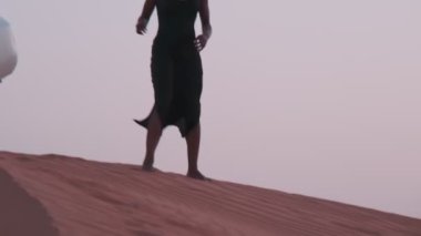 Uzun siyah elbiseli esmer bir genç kadın ve bir Arap atkısı çöl kumunda çıplak ayakla kumsalda duran beyaz bir SUV 'ye doğru yürüyor. Yavaş çekim