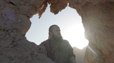 Genç sakallı bir şaman çölün kum tepelerinin arasında bir kaya mağarasında dururken dua eder. Yavaş çekim
