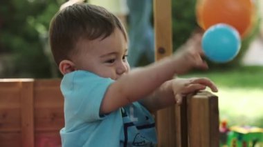 Küçük bir çocuk ve annesi çimenlikteki ahşap bir kuşhanede renkli toplarla oynuyorlar. Yaz, güneşli bir gün. Yavaş çekim, Yakın çekim.