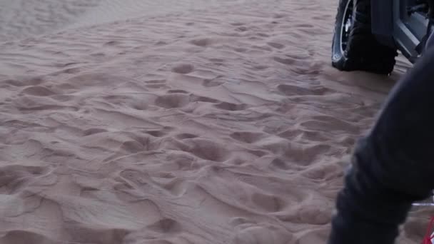 スポーツウェアの若い女性は砂漠の砂の上を歩き 夕日を背景に砂丘の上に立っているバギーに歩きます スローモティボ — ストック動画