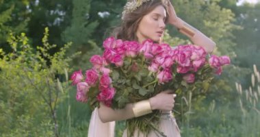 A young girl with a wreath of flowers holds a large bouquet of roses among the spring garden. Slow motion