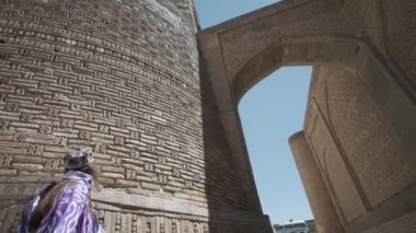 Dalgalı saçlı, uzun doğulu cübbesi ve kafatası şapkası olan genç bir kadın, minarenin önündeki meydanda yürüyor. Yavaş Hareket, Özbekistan.
