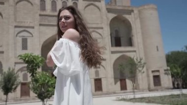 Açık beyaz elbiseli ve sandaletli, dalgalı saçlı genç bir kadın antik bir doğu şehrinde yürüyor. Yavaş Hareket, Özbekistan