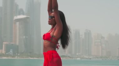 Bikinili esmer ve açık kırmızı elbiseli genç bir kadın denizin ve Dubai 'nin gökdelenlerinin önünde çıplak ayakla kayalarda yürüyor. BAE, Ağır çekim.