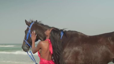 Kırmızı elbiseli genç esmer bir kadın okyanus kıyısında bir atın yanında duruyor. Yavaş çekim.