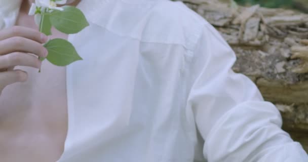 手に白い花を持つ薄い白い服を着た若い男が緑の開花庭の中に座っています スロームーン — ストック動画