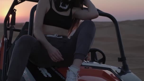 スポーツウェアを着た若い女性は 日没を背景に砂漠の真ん中のバギーに座っています スローモーション — ストック動画
