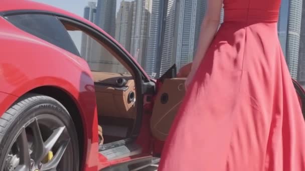 一辆昂贵的红色跑车在迪拜海滩的沙滩上缓缓行驶 旁边是一位头发飘扬 身穿红色长裙的年轻女子 慢动作 — 图库视频影像