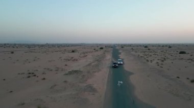 Bir İHA çölün kumları arasında boş bir yolda giden iki pahalı arabanın yanından uçuyor. Hava Görünümü, Yavaş Hareket.