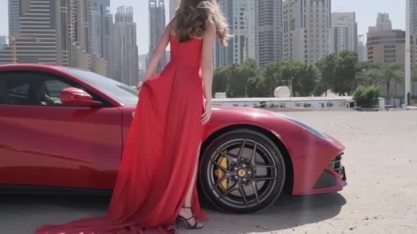ドバイの高層ビルを背景に高価なスポーツカーの隣には 長いドレスとハイヒールを履いた若い女性が立っています スローモーション — ストック動画