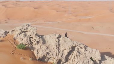 Bir dron, Birleşik Arap Emirlikleri 'nin kumlu çölündeki bir uçurumda dua eden şaman kılığında sakallı bir adamın etrafında uçuyor. Hava görünümü