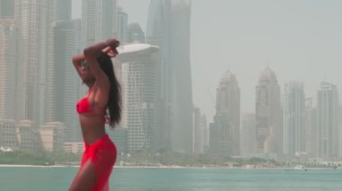 Bikinili esmer ve açık kırmızı elbiseli genç bir kadın denizin ve Dubai 'nin gökdelenlerinin önünde çıplak ayakla kayalarda yürüyor. BAE, Ağır çekim.