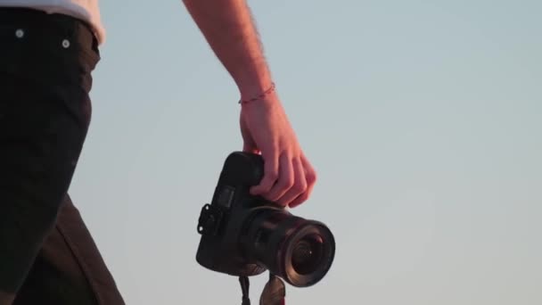 彼の手にカメラを持った若い写真家が日没の光の中で砂漠の砂の上を裸足で歩く スローモーション — ストック動画
