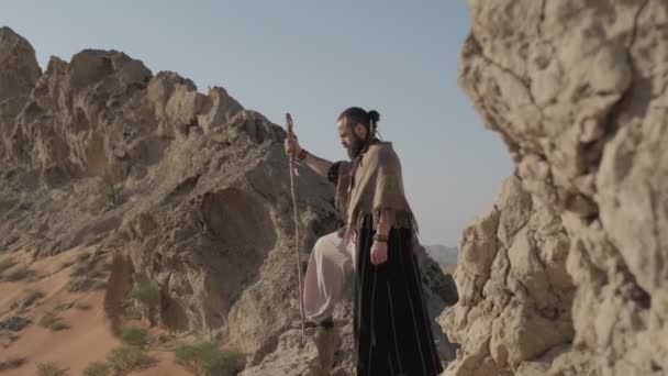 砂漠の真ん中 澄んだ空に向かって孤独な岩の上に スタッフを手にシャーマンの格好をした若い髭を生やした男が立っている スローモーション — ストック動画