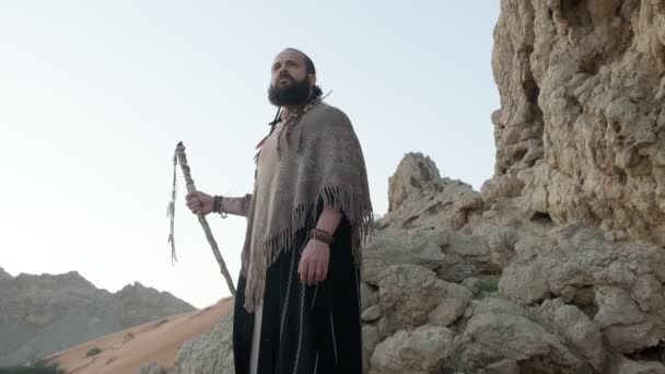 砂漠の真ん中の岩の上を若い髭を生やしたシャーマンが歩いている スローモーション アラブ首長国連邦 — ストック動画